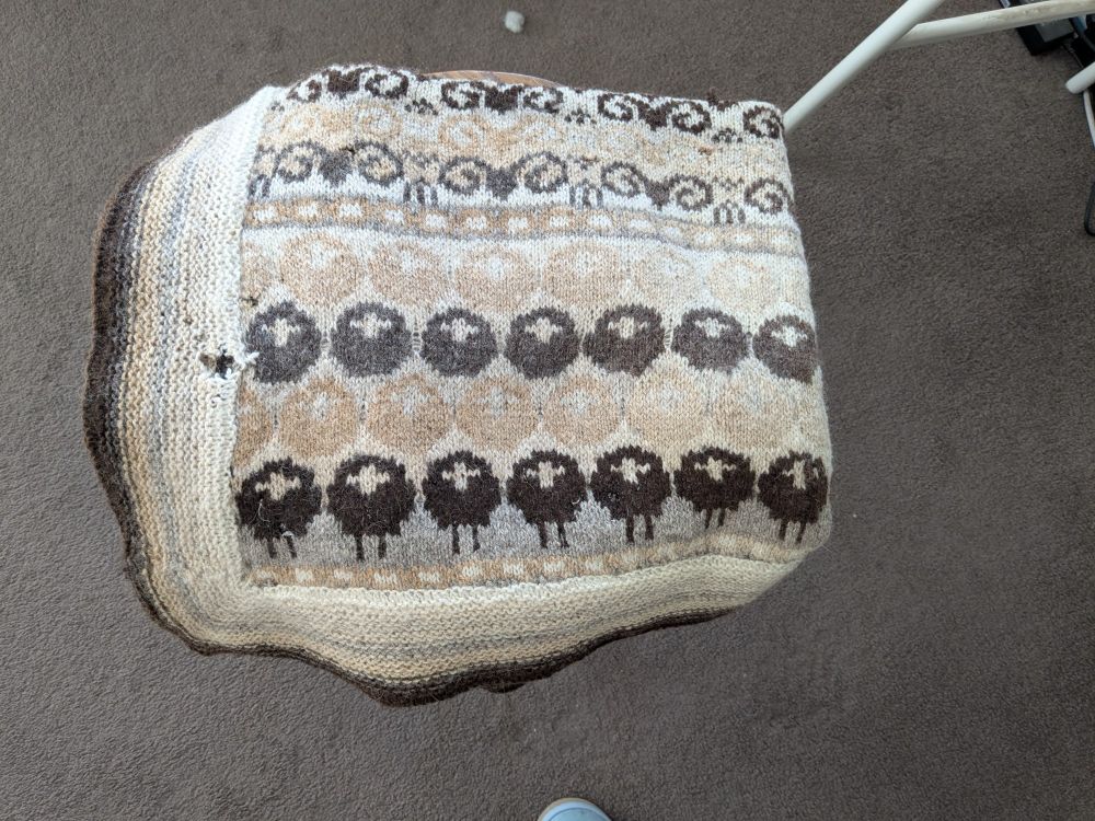 A blanket, knitted in natural wool colours with sheep motives. It has holes, large and small.