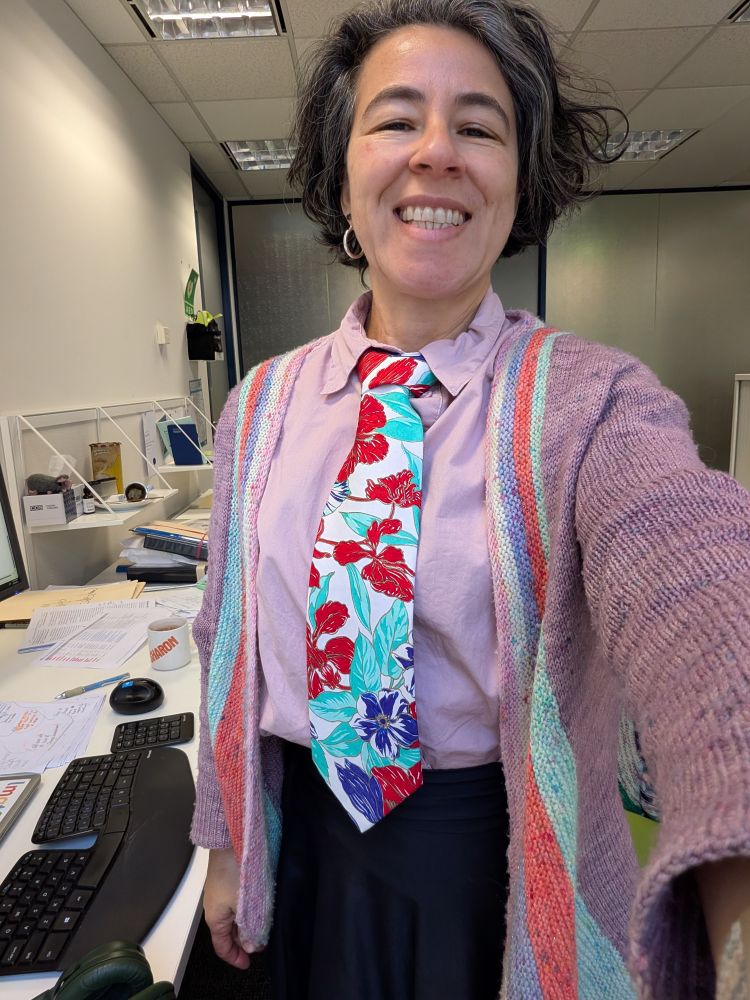 A woman in a light purple shirt and cardigan wears an aggressively floral patterned turquoise red and navy tie.