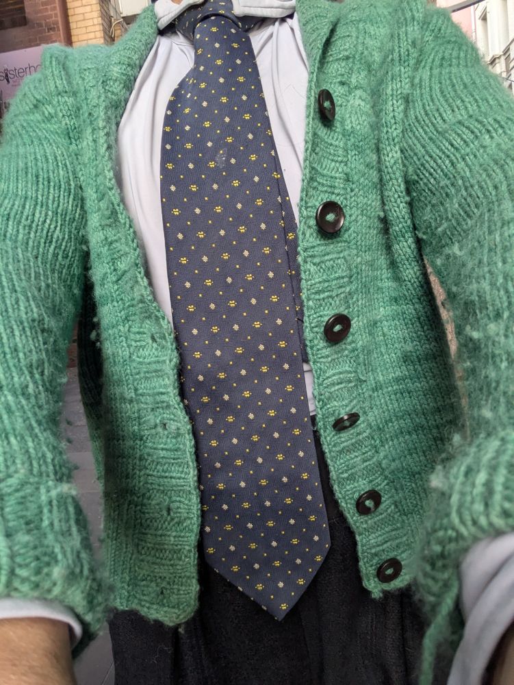 A woman wears a green cardigan and a navy tie with a small gold repeating pattern