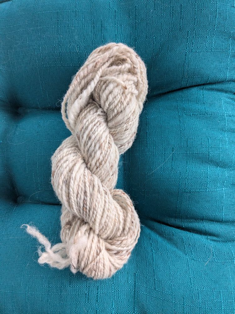 A 50 gram skein of oatmeal coloured handspun yarn sits on a teal cushion.