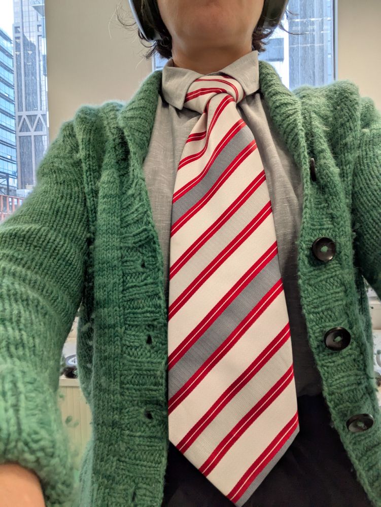 A woman wears a wide tie. The tie has diagonal stripes, thin red, thick in white, light grey and dark grey.