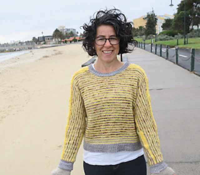 A woman wearing a yellow and grey striped jumper stands at a beach. It's windy and she is struggling to smile.