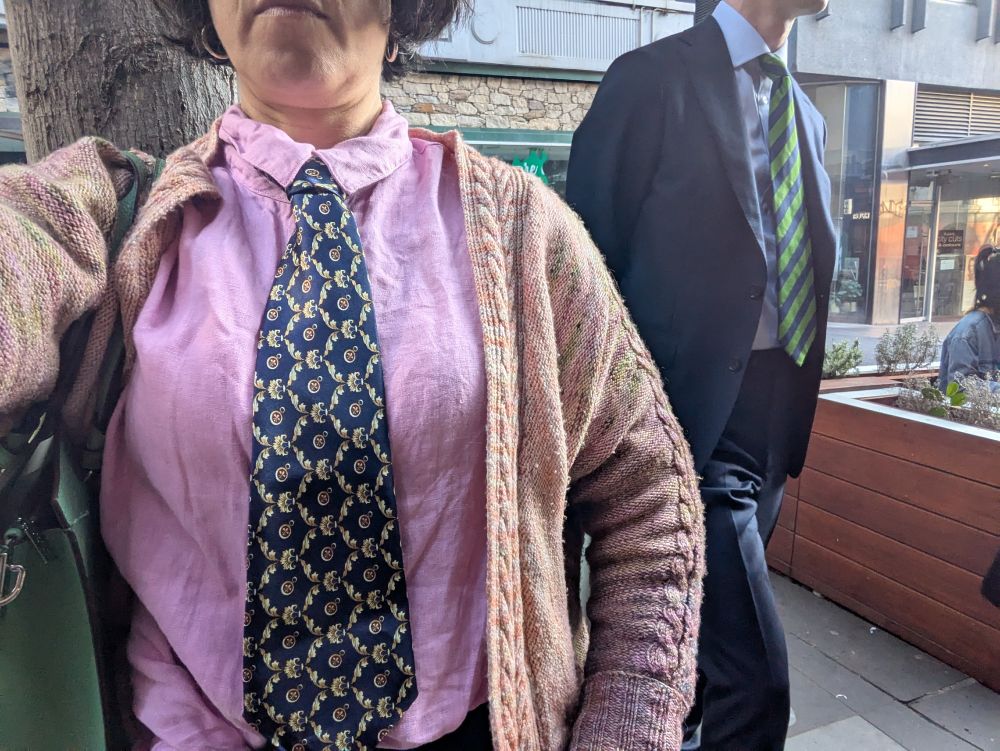 A woman in a pink shirt wears a tie with a geometric pattern. Behind her is a man wearing a wrinkled tie with diagonal stripes. You cannot see either of their faces.