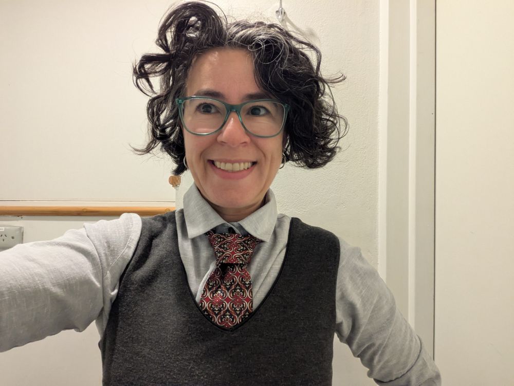 A woman with a manic smile. She is wearing the same tie as in the previous photo, but now she has a great tunic dress on.