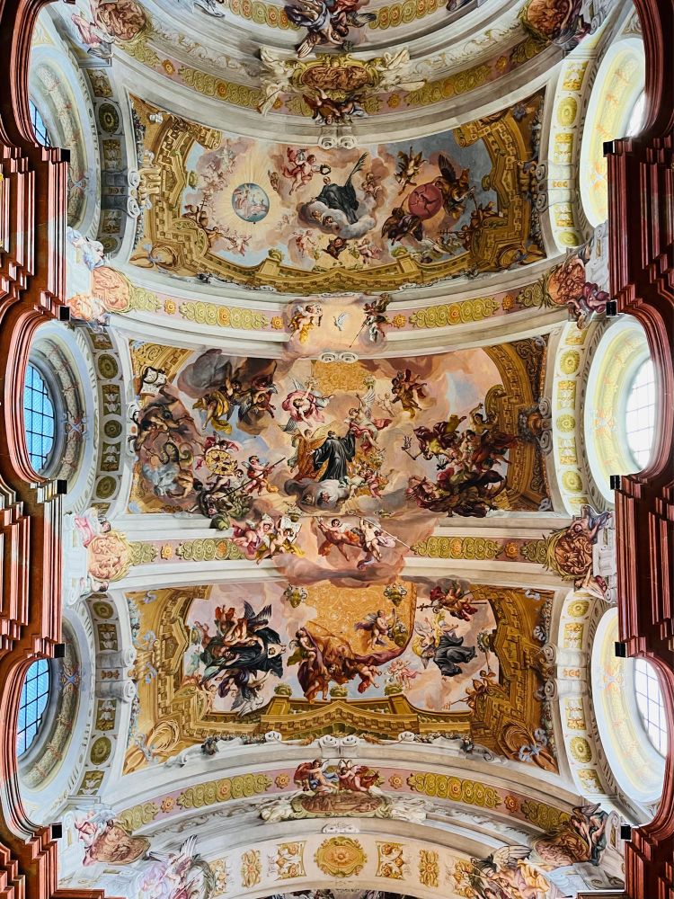 Farbfoto (Hochformat)
Der Blick richtet sich auf das großflächige, polychrome Decken- und Kuppelfresko im Stil des Hochbarock: Zwischen den elliptischen Obergadenfestern („Ochsenaugen“) öffnet sich ein „exaltierter Himmel“, in dem illusionistische Architektur und extrovertierte Heiligenverehrung die Symmetrielinie nachvollziehen. Es überwiegen Gelb-, Ocker- und Rottöne, die das Weiß der Quergurte konterkarieren.
#StiftMelk