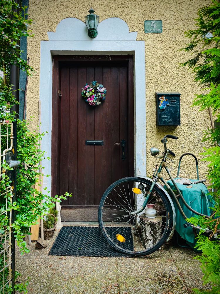 Farbfoto (Hochformat)
Kleiner Vorgarten hinterm Zaun: Blick auf eine Haustüre, links davon lehnt ein Fahrrad. An der Haustüre hängt ein bunter Blumenkranz.