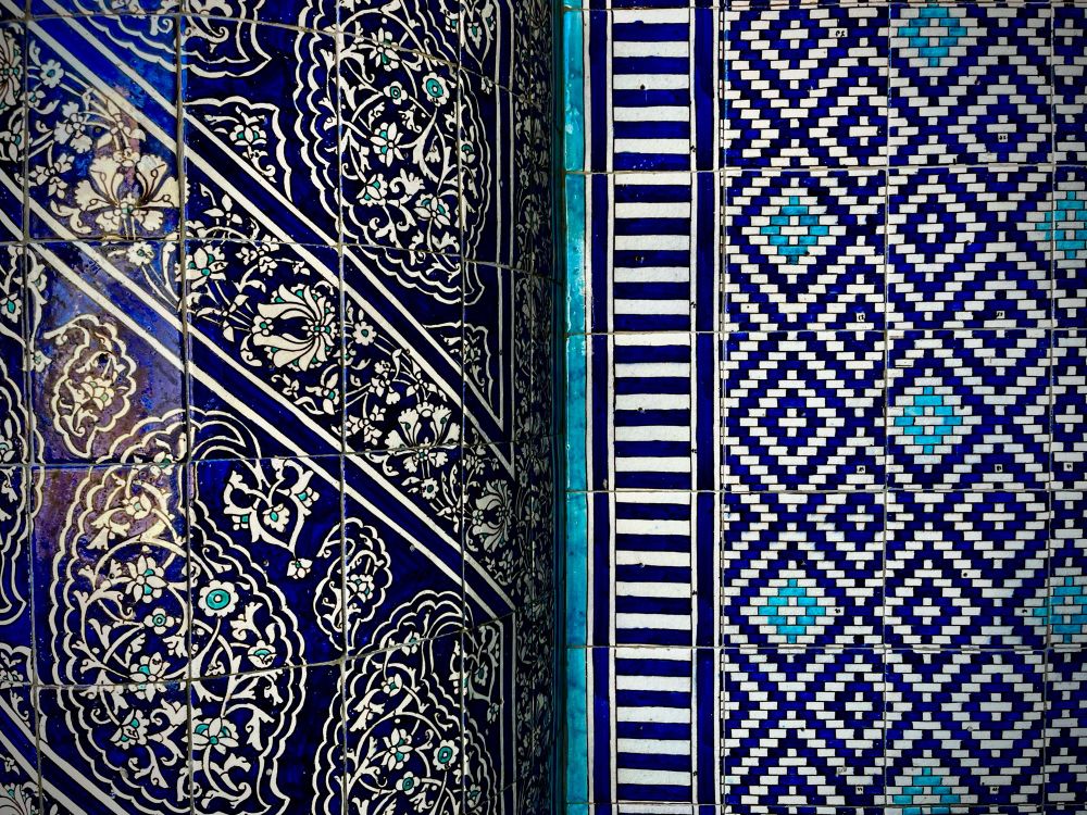 Farbfoto (Querformat)
Wanddekoration - Keramik-Fayencen in Kobaltblau und Weiß: Rechts, auf planem Untergrund, eine geometrische Anordnung von kleinen weißen und blauen Mosaiksteinchen, die ein regelmäßiges Rautenmuster bilden. In der linken Bildhälfte der Ausschnitt einer angrenzenden Halbsäule, die mit dekorativen, blauweißen Kacheln mit Blumenmotiven und Arabesken gestaltet ist. Die Bildmitte bildet ein horizontaler Strich in Türkis.