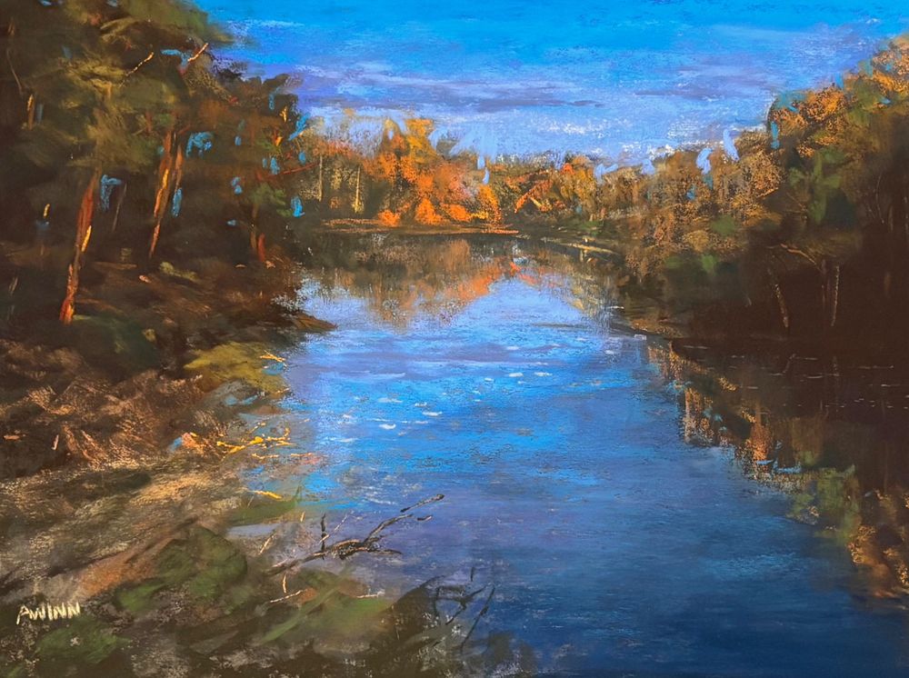 A 9x12” soft pastel painting. Tree lines surround a pond that is receding towards the horizon line. The pond reflects the blue and purple sky. The trees are a mix of green and orange foliage. 