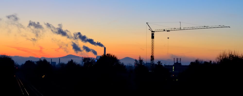 Vor einem Sonnenaufganghimmel zieht in der linken Bildhälfte Rauch aus zwei Industriekaminen nach links und in der rechten Bildhälfte ist ein großer Baukran mit seinem Ausleger nach rechts geschwenkt.