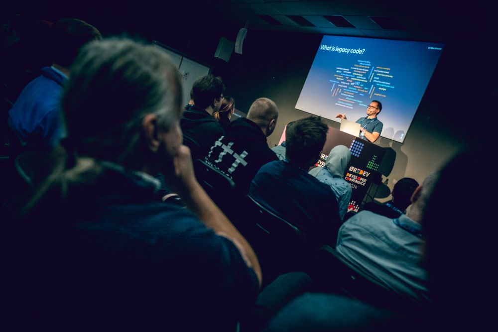 A room full of developers listening to a tech talk. The presentation shows the question What is legacy code, with a wordcloud beneath it.