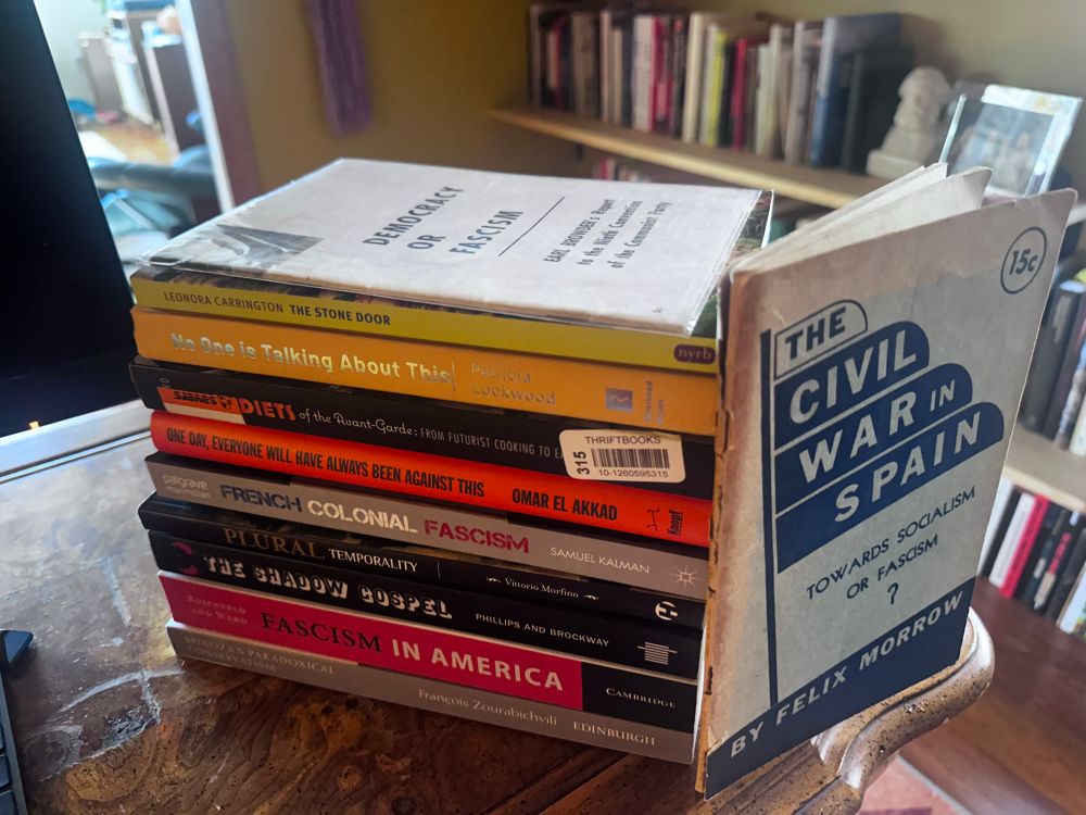 Stack of books on a wooden table, titles of:
Democracy or Fascism, Earl Browder
The Civil War in Spain, Felix Morrow
The Stone Door, Leonora Carrington
No One is Talking About This, Patricia Lockwood
Antidiets of the Avant-Garde, Cecilia Novero
One Day, Everyone Will Have Always Been Against This, Omar El Akkad
Plural Temporality, Vittorio Morfino
The Shadow Gospel, Phillips and Brockway
Fascism in America, Rosenfeld and Ward
Spinoza’s Paradoxical Conservatism, François Zourabichvili