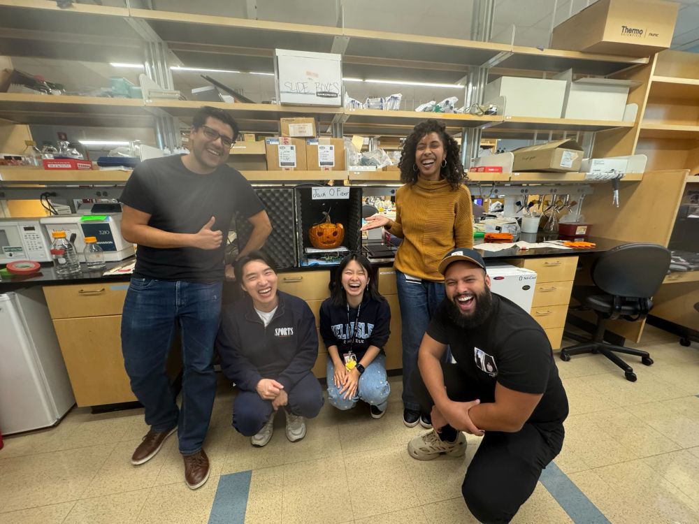 Brielle is standing in front of the lab bench with four other team members laughing. They're standing around a pumpkin carving inside of a behavior box. 