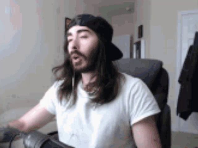 a man with long hair and a beard is sitting in a chair wearing a hat and a white shirt .