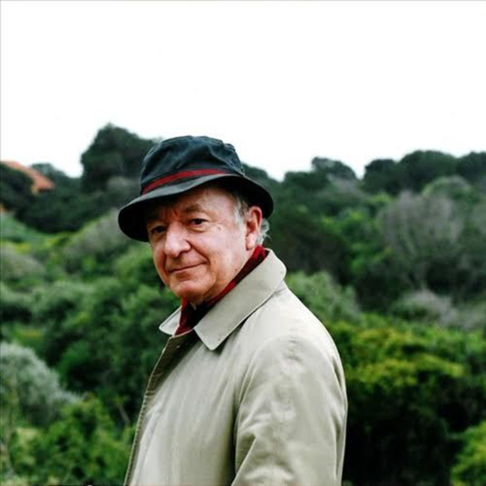An older man wearing a green hat and a trench coat standing in front of a tree covered hill