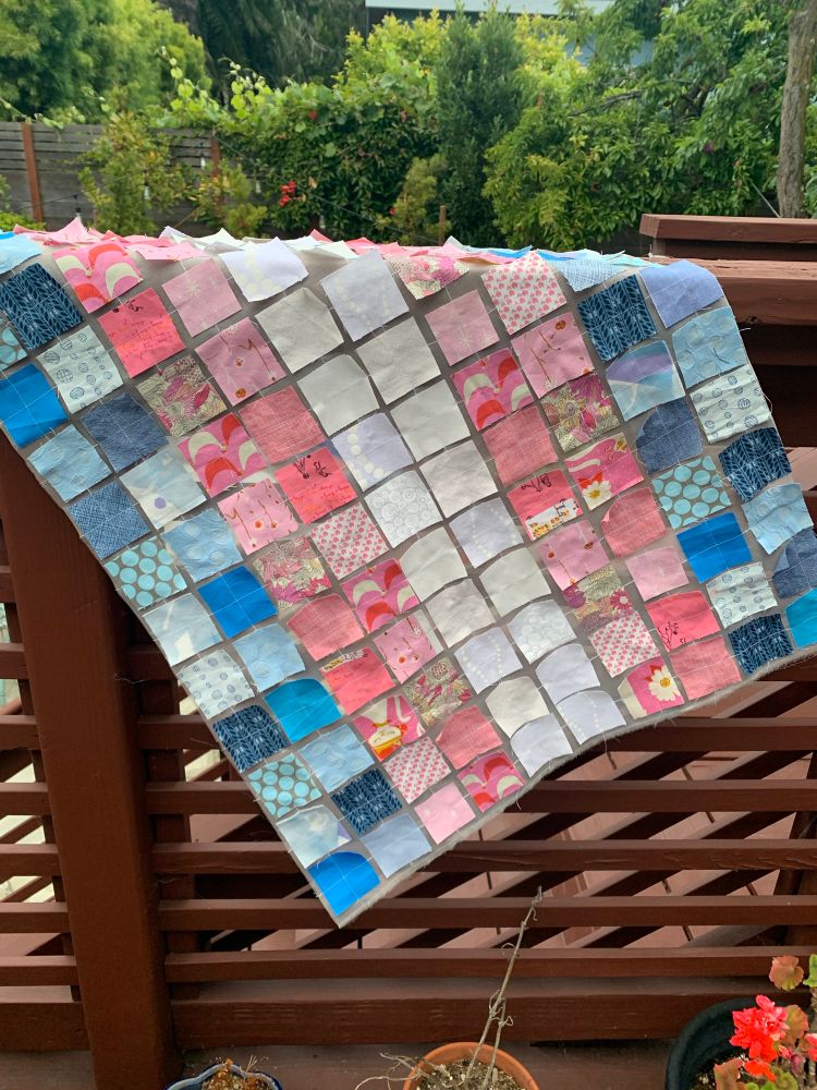 Homemade fabric trans flag made of squares of quilting cotton in shades of light blue, light pink, and white, tacked on to a sheet of white silk organza. The flag is draped over the brown wooden banister of a deck.