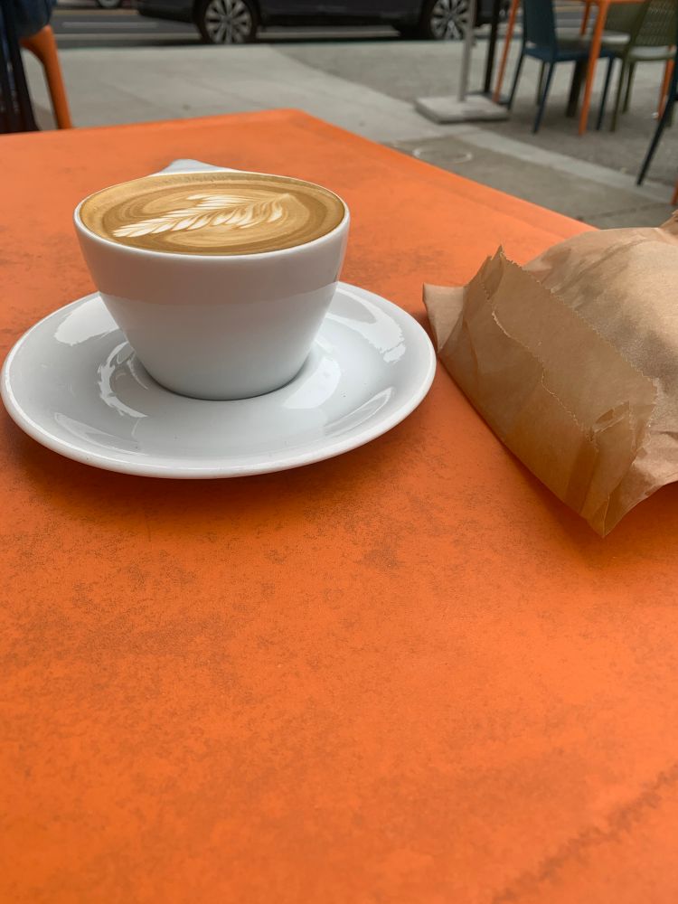 A cappuccino in a white teacup with saucer and a small brown paper bag on a dirty orange plastic table at a sidewalk cafe 