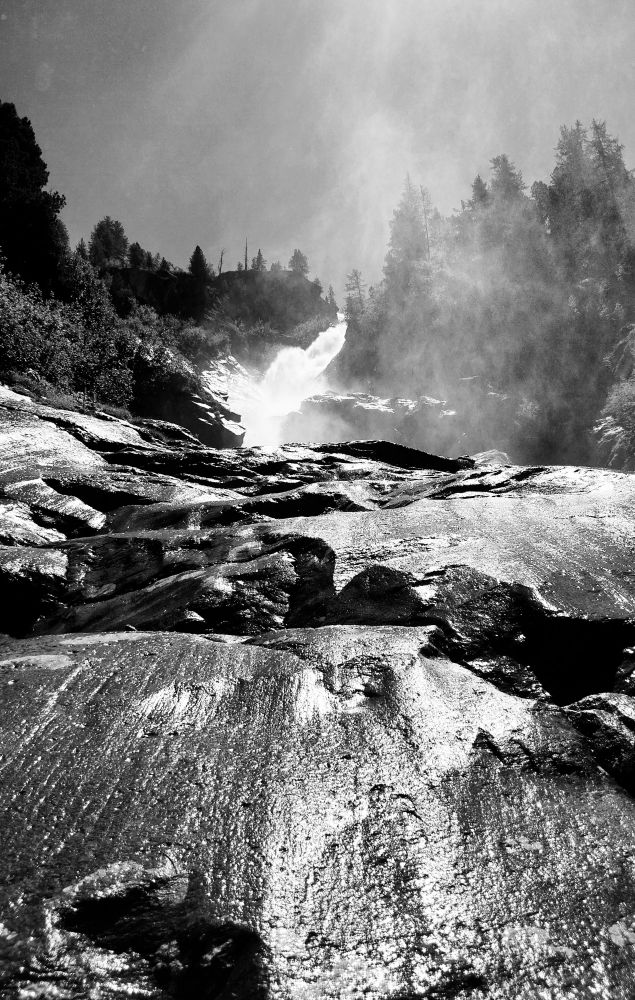 A waterfall seen from below, with the wet rocks eroded by the water in the foreground. Above, the mass of water cascading from a coniferous forest and the water vapor. 