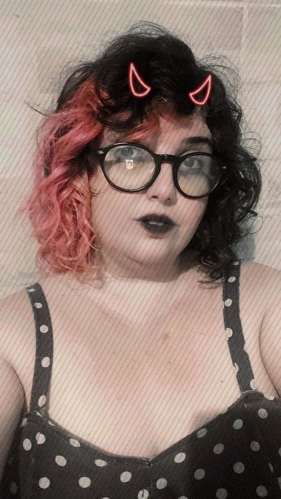 Uma mulher branca, de batom preto, cabelos metade pretos, metade rosa e de chifrinhos. Veste um vestido preto de bolinhas.