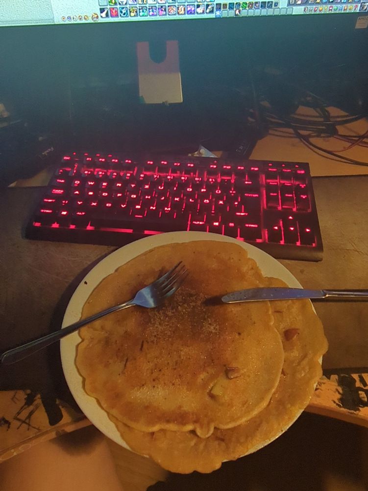 Ein Teller auf einem unaufgeräumten, mit Kabel und Computerbauteilen verwüsteten Schreibtisch, bedeckt mit frischen Pfannkuchen mit Apfelstücken, mit Zimt und Zucker bestreut.