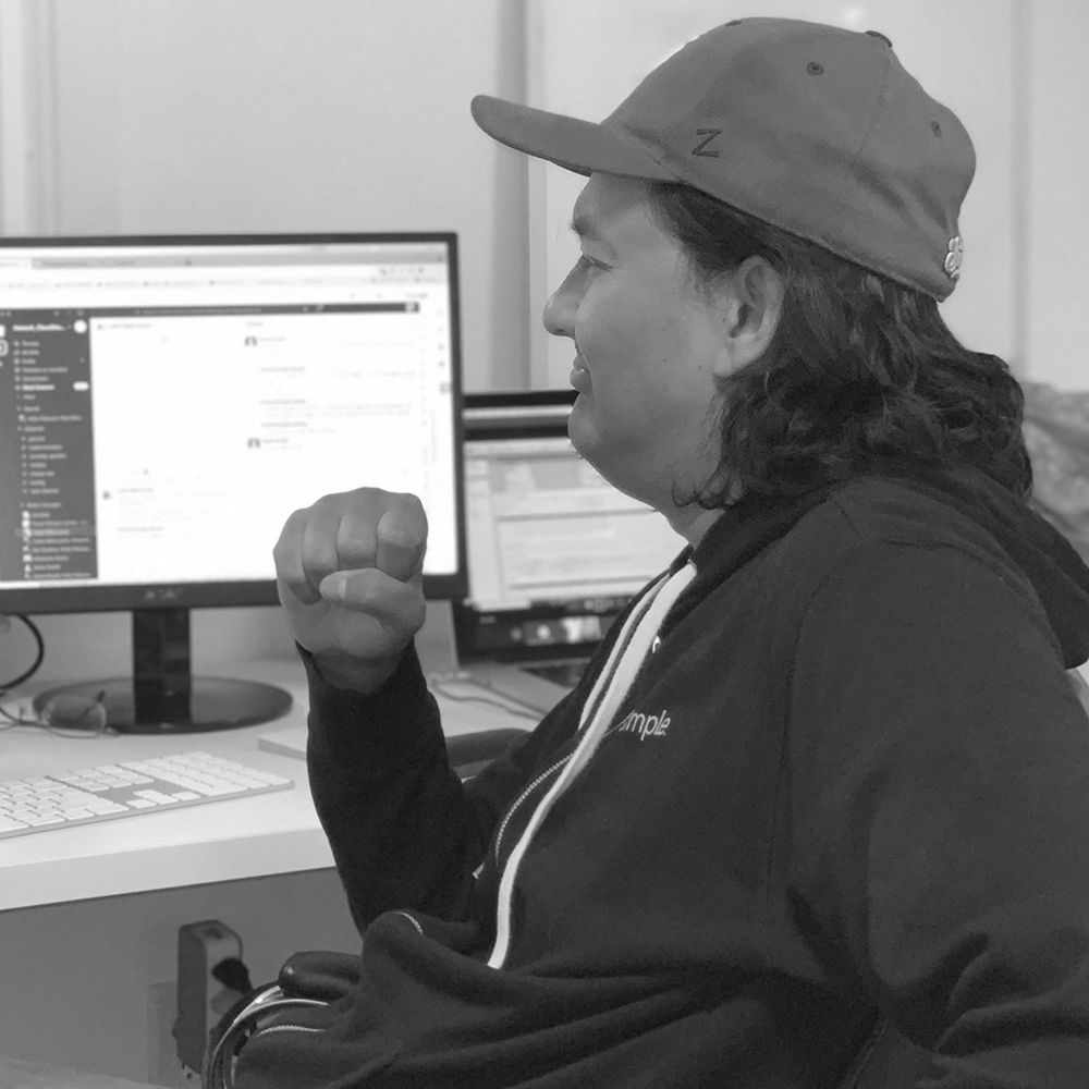 A man sits in front of several computer screens  while looking at them with a serious look on his face. Angle is from the side. He is wearing a baseball cap & hoodie.