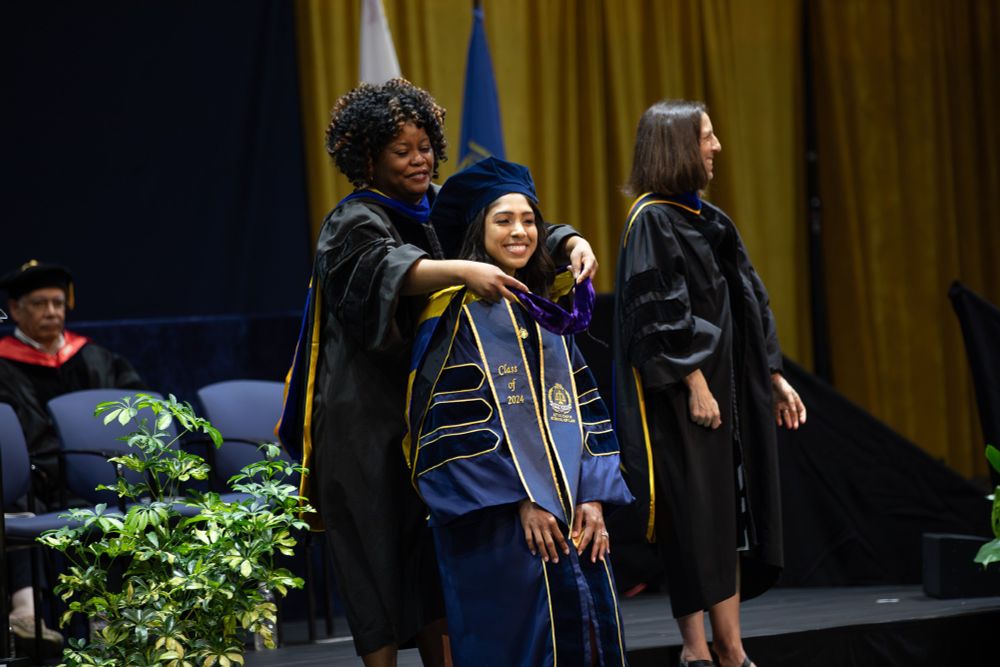 A graduate wearing their regalia has their hood placed on their shoulders during the commencement ceremony. 