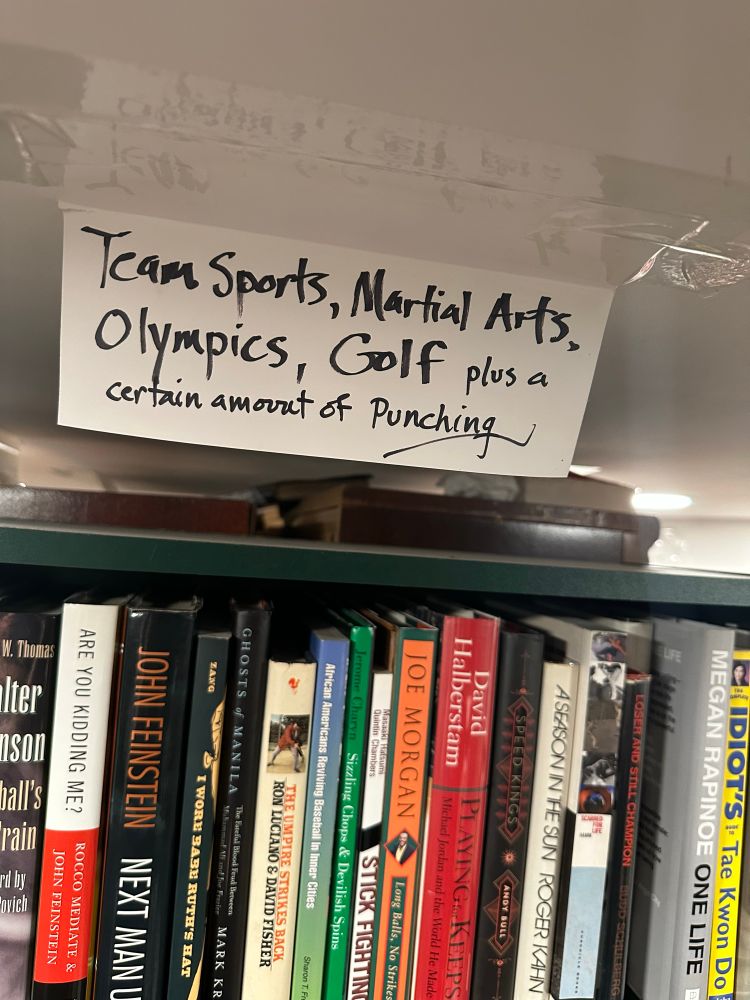 A shelf of used non-fiction sports-themed books with a hand written shelf tag “team sports, martial arts, Olympics, golf, plus a certain amount of punching.”