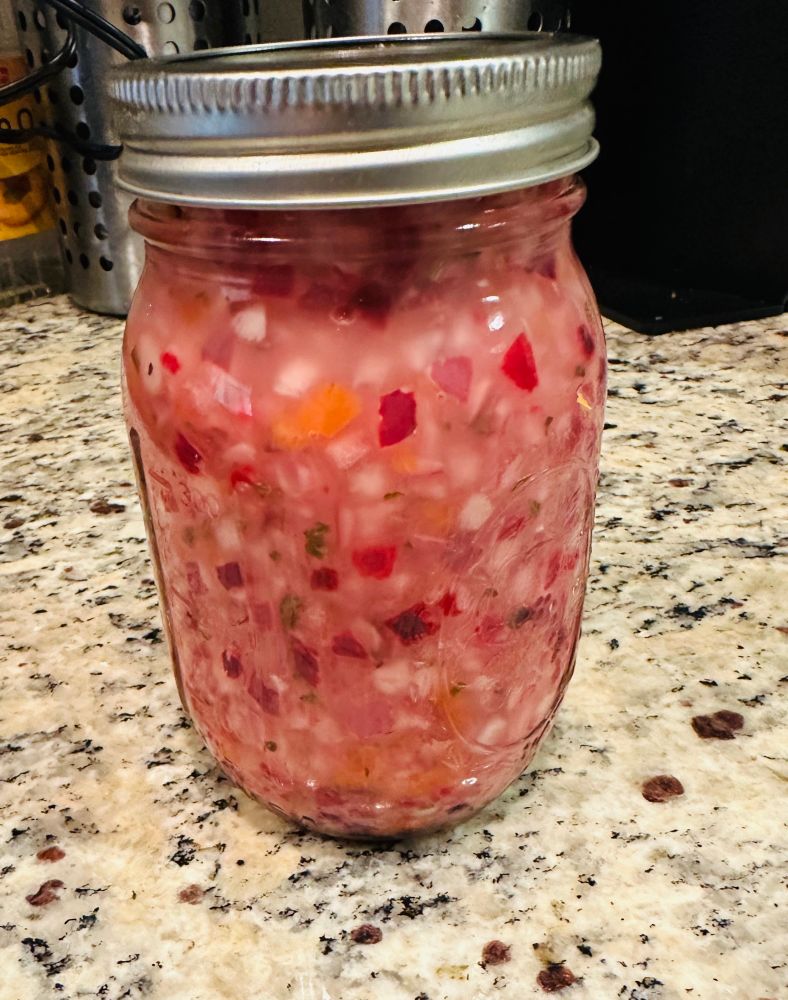 A mason jar sits on a stone countertop. The jar is filled with a bright pink mix of diced red onion and habanero peppers, like party confetti for your taste buds.