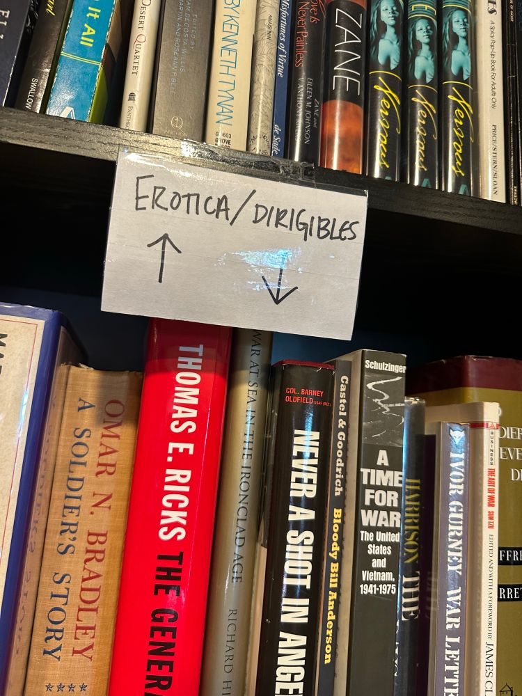 A tag between two shelves of used books, with an up arrow next to “Erotica” and a down arrow next to “dirigibles,” and possibly never have those two topics been grouped together outside of dirigible smut, which I’m sure is an actual subgenre somewhere.