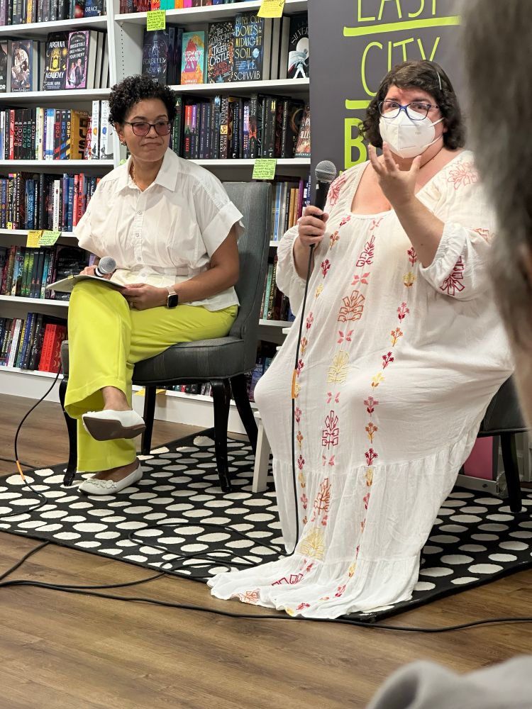 Mia Sosa and Olivia Dade seated together interview-style in front of a wall of books at East City Bookshop. Mia is Latina with short curly hair, dark glasses, bright cheerful spring green/yellow slacks, and a breezy white blouse. Olivia is a fat white woman with  straight, dark hair pulled back with cute barrettes, and a long white linen dress beautifully embroidered in pink, red, and yellow.
