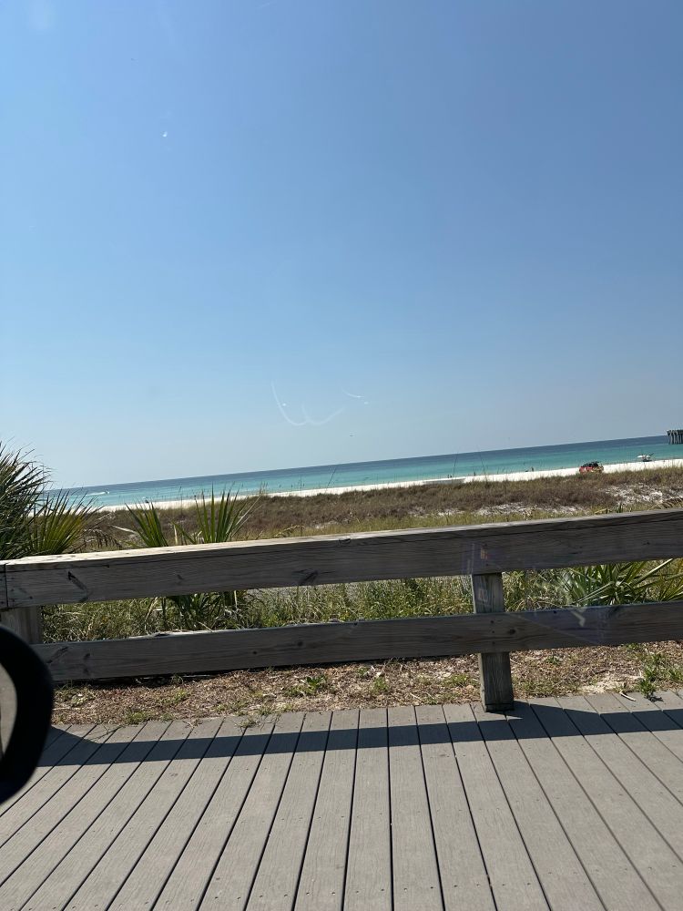 A perfectly calm, crystal blue Gulf of Mexico beneath a clear sky viewed from a boardwalk. Grassy dunes and white sand beaches lay between the boardwalk and the water.