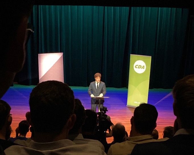 CDA-leider Henri Bontenbal staat midden op het podium aan de voorkant van de theaterzaal. Hij draagt een marineblauw pak en naast hem staat aan elke kant een banner, eentje van het CDJA en eentje van het CDA.