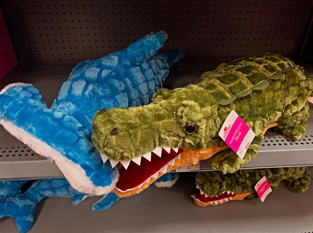 A plush alligator is chomping down on the head of a plush hammerhead shark. Both are labeled as Valentine's plush toys, and are displayed in the Valentine's Seasonal department at Walmart. 