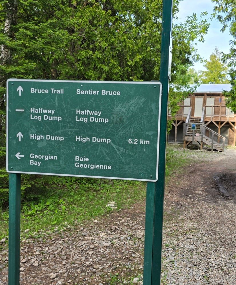 A Parks Canada directional sign. It indicates that high dump is 6.2 km in that way.  Immediately in the direction that arrow points of a vaulted toilets building.