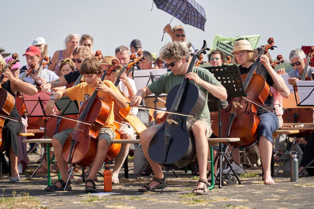 sehr viele Celli spielen auf dem Tempelhofer Feld, im Vordergrund erkennbar ein kleiner Junge und ein Mann mit einem schwarzen Cello