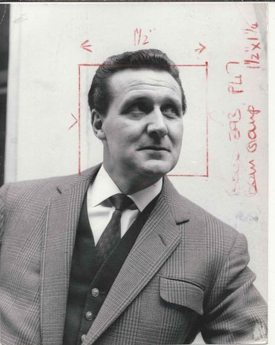 Black and white photo of Patrick MacNee standing in front of a red diagram on white paper.
