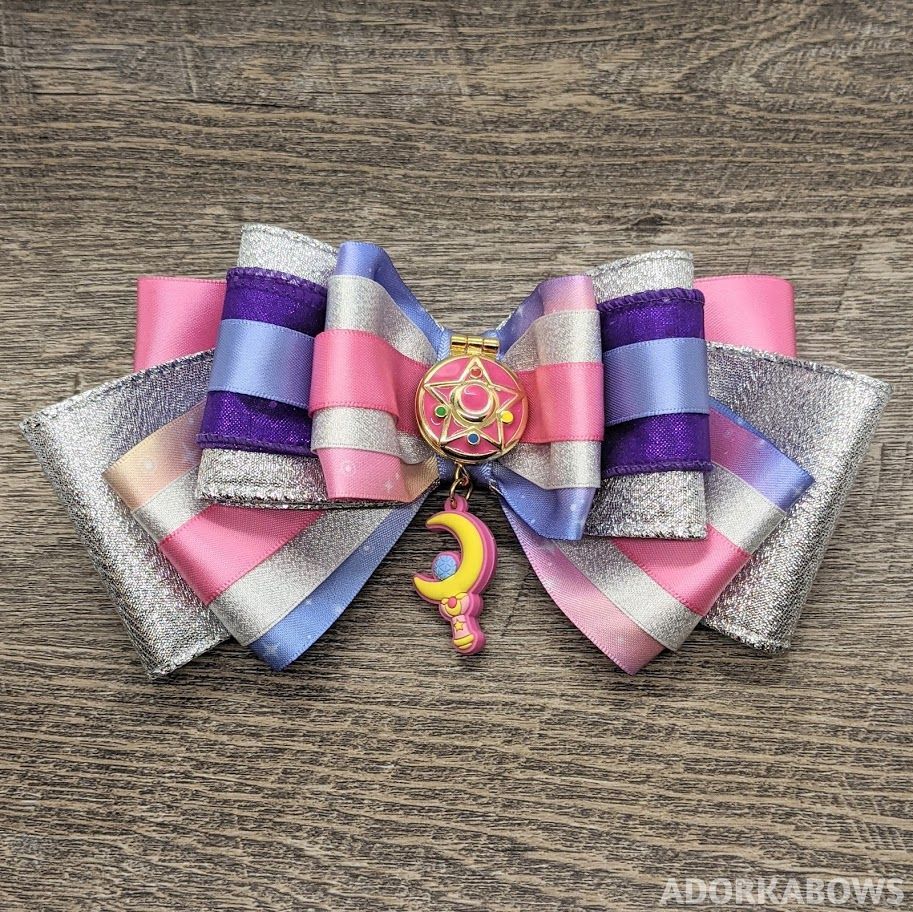 Silver, pink, and purple bow with a Sailor Moon Brooch in the center, a small vinyl moon wand hanging from it.

