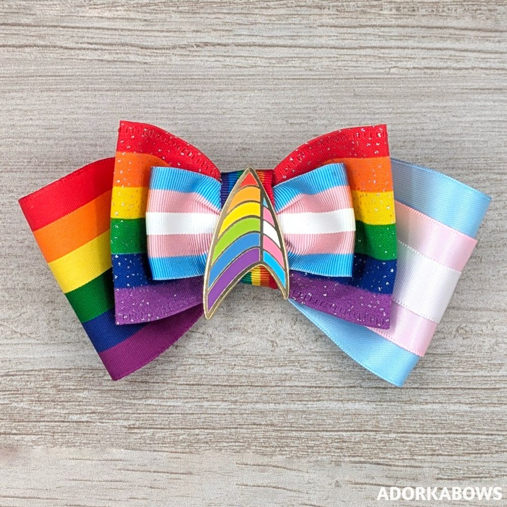 A hair bow in rainbow and trans Pride colors with a center pin shaped like the Star Trek uniform emblem in rainbow and trans Pride colors