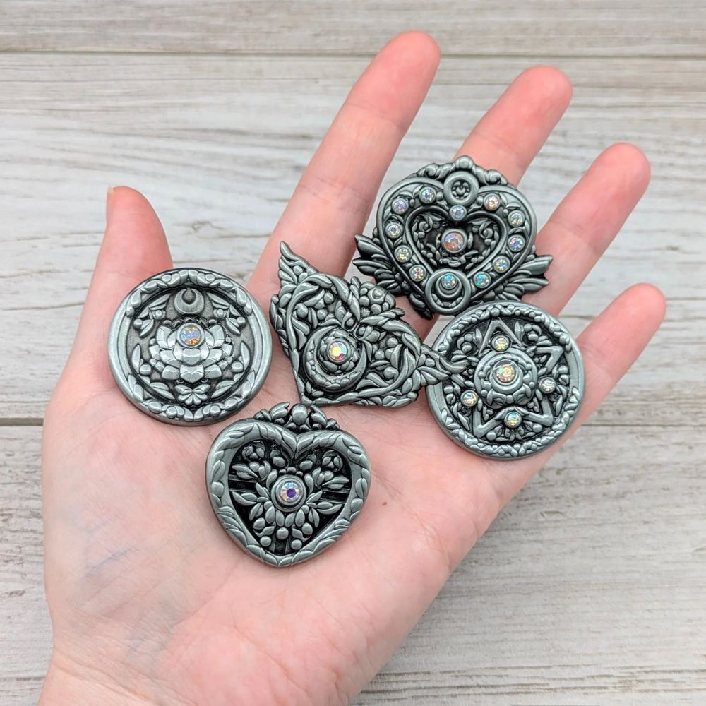 A hand holding antique silver metal pins in the shape of Sailor Moon brooches. There are five of them. Pins by Adorkabows & AmunetGFX.