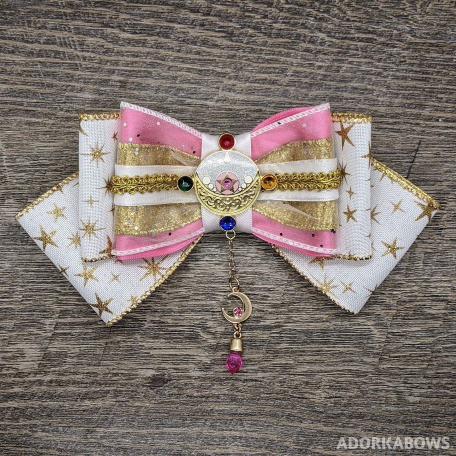 Pink, white, and gold bow with a Sailor Moon brooch in the center, a gold moon dangling from it.