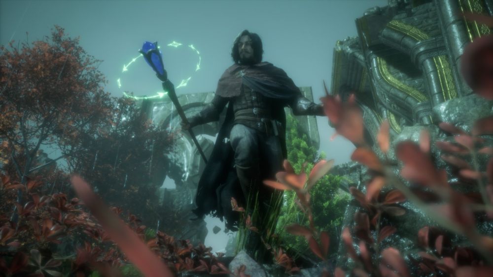 A digital photograph from Dragon Age The Veilguard. It shows the subject, a white older man with medium-length greyish brown curly hair with a mustache and goatee. He is in tattered adventurers attire while wielding a staff, which has green magic circling the blue jewel on the tip of the staff. Red leaves frame the bottom foreground while greyish green skies loom overhead and old stone ruins in the shape of arches frame the background of the shot.