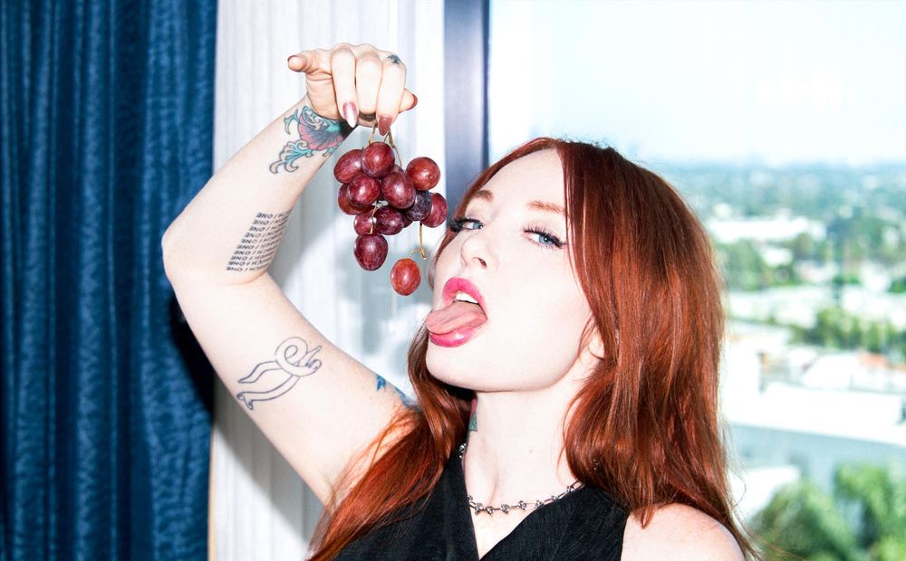 A redhead woman is sticking out her tongue, holding a bunch of grapes over her open mouth as if to eat one.