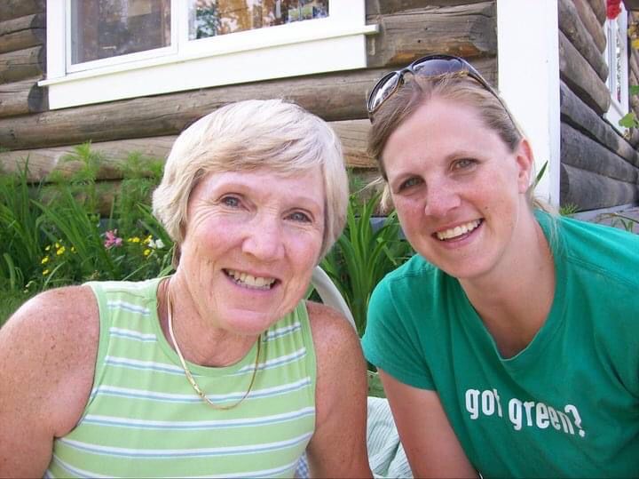 Woman and her adult daughter smile together in front of an old log cabin.