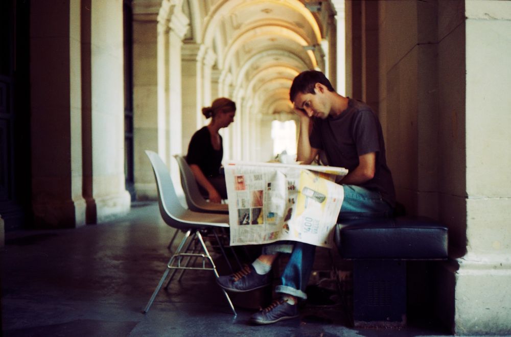 Man seated at cafe table reads broadsheet newspaper