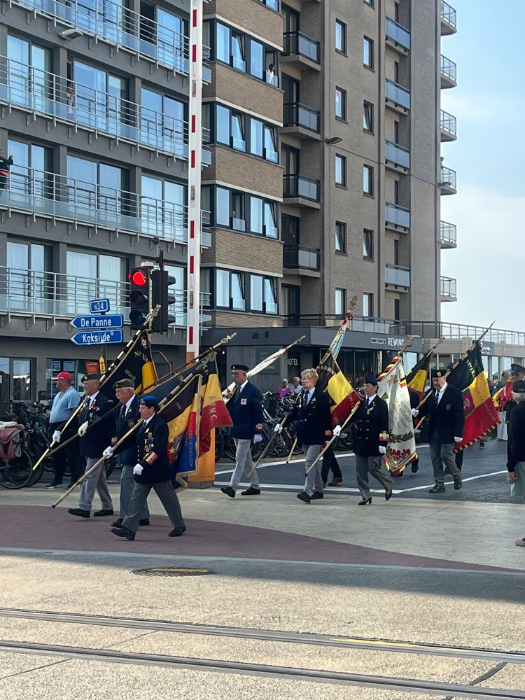 Belgian military dignitaries in uniform bearing  black-yellow-red flags and walking along a street in Oostduinkerke. There’s an apartment building and a traffic light in the background. 