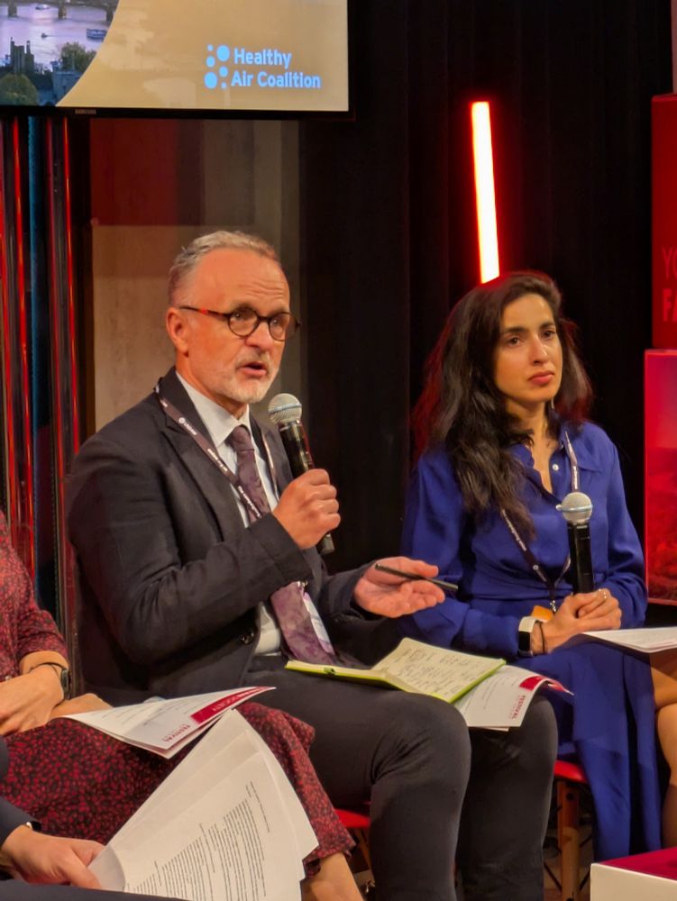Dr Mike McKean sits on a stage next to the Chair and other panellists, and speaks into a microphone.