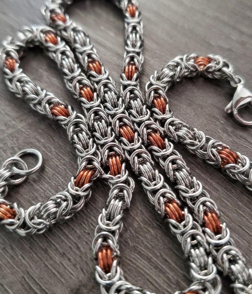 a close up of a slim chain necklace on top of a gray wood desktop. it is made with the byzantine chainmaille weave using mostly stainless steel rings, and features copper rings at evenly spaced intervals in the weave (at every fifth step of the byzantine units).
