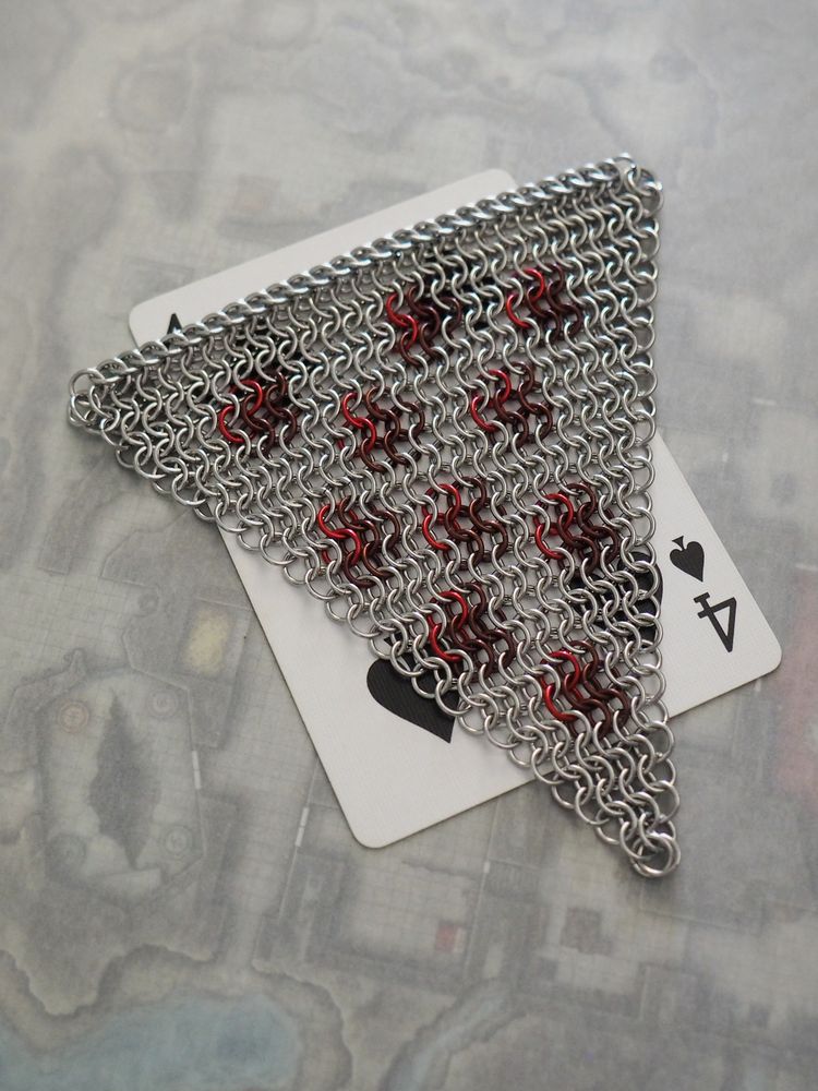 a triangle shaped chainmaille fidget toy sits on top of a four of spades playing card to show size (just a bit longer and wider). the fidget is made mostly with stainless steel, paired with anodized aluminum for the pepperoni slices. it's in the classic euro 4 in 1 pattern, but with a 3 in 1 half persian weave at the crust.