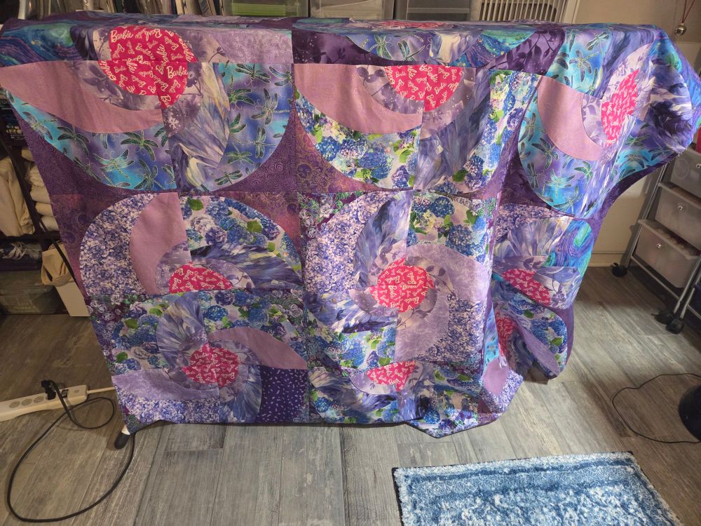 About half of a quilt top draped over an ironing board.  Colors are lavender, purple,  blues, and greens with Barbie pink centers. Gray floor,  part of a blue printed rug in the lower right of the picture.  