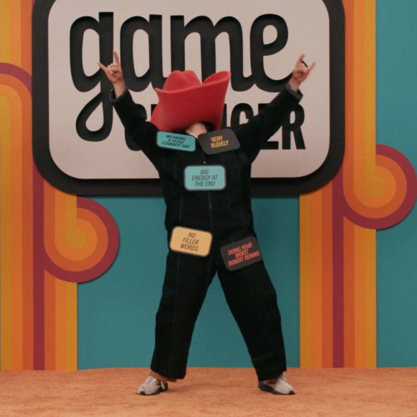 screenshot of anna garcia from game changer in a black jumpsuit with a giant red cowboy hat covering their head. they have cards stuck all over their body and are holding up their hands in a sign of the horns/rock hands