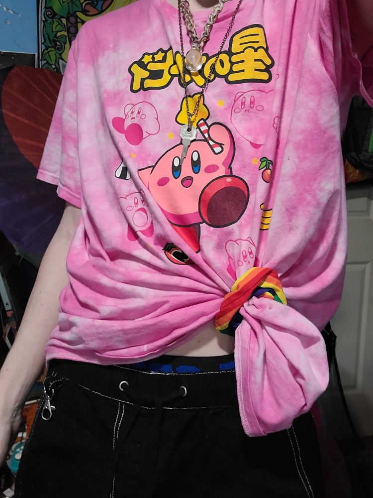 Dart showing his outfit of the day, a pink tie dye kirby t-shirt tied with a rainbow scrunchie to the left. Black shorts trimmed with white stitched show off the top of his black and blue cellblock13 jockstrap. He's also wearing 2 necklaces....one of which is to his cage he's currently wearing.